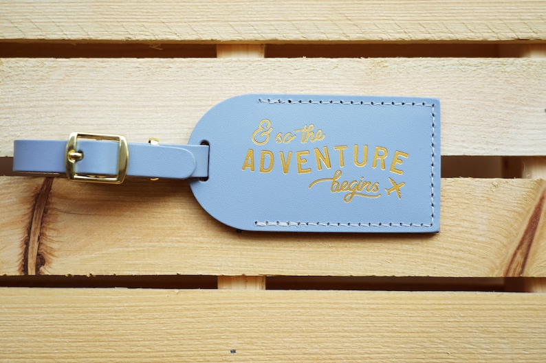 May include: A light blue leather luggage tag with gold stitching and the text "& so the adventure begins" in gold lettering. A gold airplane icon is next to the text.
