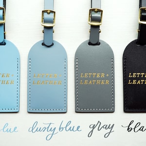 K&ouml;nnte beinhalten: Vier Leder-Gep&auml;ckanh&auml;nger in Himmelblau, Staubblau, Grau und Schwarz mit goldener Beschriftung "LETTER + LEATHER".