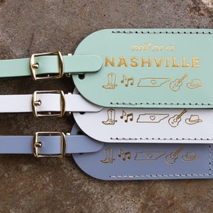 May include: Three leather luggage tags in mint green, white, and gray with gold stitching. Each tag has the text "meet me in NASHVILLE" and gold icons of a boot, musical notes, the state of Tennessee, a guitar, and a cowboy hat.