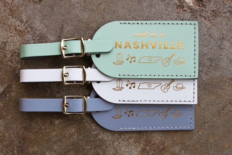 May include: Three leather luggage tags in mint green, white, and gray with gold stitching. Each tag has the text "meet me in NASHVILLE" and gold icons of a boot, musical notes, the state of Tennessee, a guitar, and a cowboy hat.