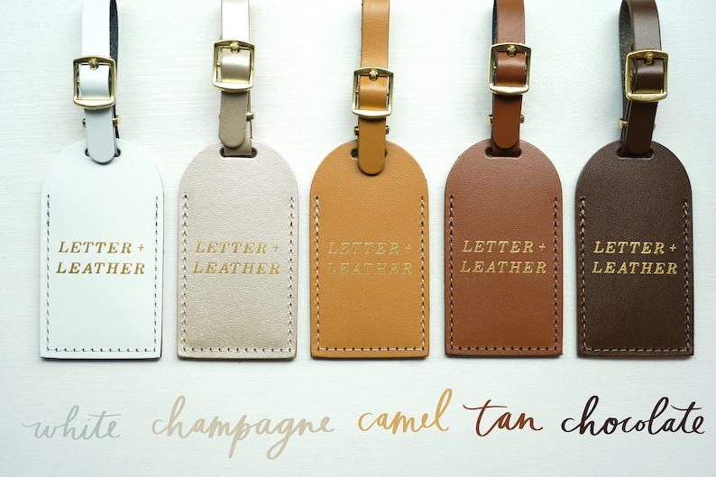 Op de afbeelding: Vijf leren bagagelabeltjes in verschillende kleuren: wit, champagne, camel, bruin en chocolade. Elk labeltje heeft de tekst "LETTER + LEATHER" in goud gegraveerd.