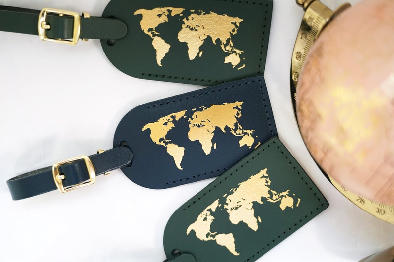 Op de afbeelding: Drie leren bagagelabeltjes met gouden wereldkaartontwerpen. Twee labeltjes zijn donkergroen en &eacute;&eacute;n is donkerblauw.