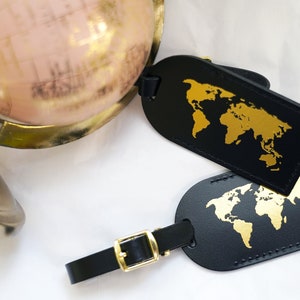 Op de afbeelding: Twee zwarte leren bagagelabeltjes met gouden wereldkaartontwerpen. De labeltjes hebben een gouden metalen gesp en een zwart bandje.