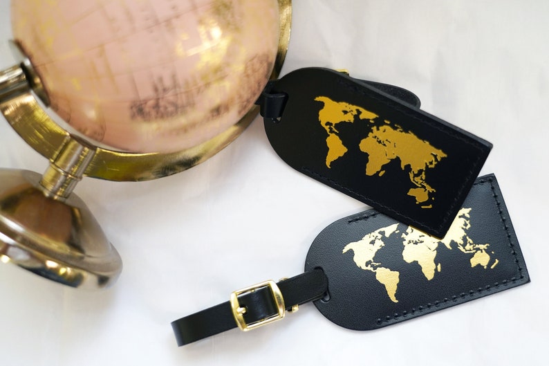Op de afbeelding: Twee zwarte leren bagagelabeltjes met gouden wereldkaartontwerpen. De labeltjes hebben een gouden metalen gesp en een zwart bandje.