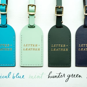 K&ouml;nnte beinhalten: Vier Leder-Gep&auml;ckanh&auml;nger in verschiedenen Farben: Tropenblau, Minze, J&auml;gergr&uuml;n und Marineblau. Jeder Anh&auml;nger tr&auml;gt den Text "LETTER + LEATHER" in Goldpr&auml;gung.