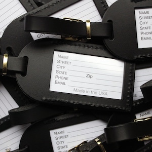 Op de afbeelding: Een close-up van een stapel zwarte leren bagagelabeltjes met witte labels. De labels hebben velden voor naam, straat, stad, staat, postcode, telefoon en e-mail. De labels zijn gemaakt in de VS.