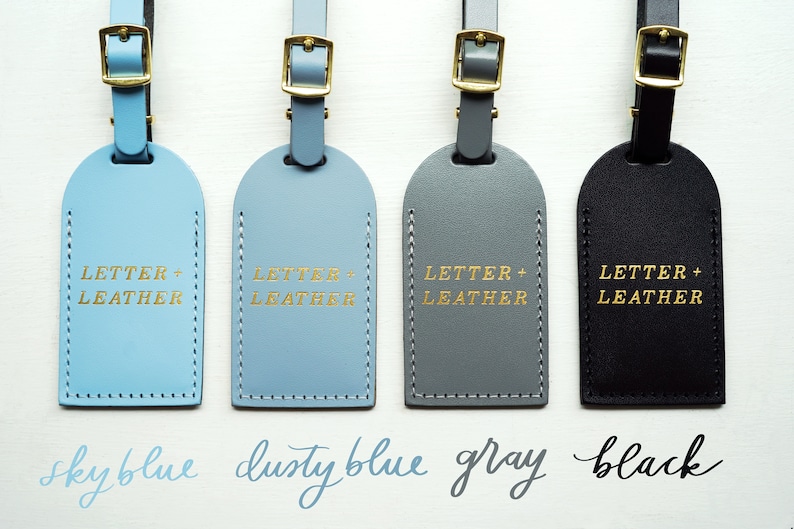 Op de afbeelding: Vier leren bagagelabeltjes in hemelsblauw, stofgrijs, grijs en zwart met gouden opschrift "LETTER + LEATHER".