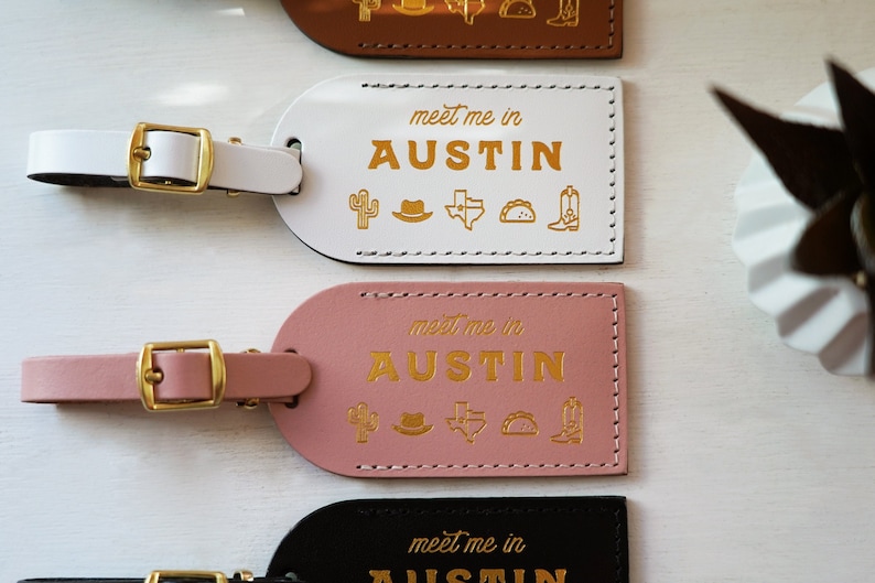 K&ouml;nnte beinhalten: Drei Leder-Gep&auml;ckanh&auml;nger mit goldenen Buchstaben und Symbolen. Die Anh&auml;nger zeigen den Text "meet me in AUSTIN" mit den Symbolen Kaktus, Cowboyhut, Texas-Staatsumriss, Taco und Cowboystiefel. Die Anh&auml;nger sind wei&szlig;, rosa und schwarz.