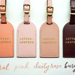 Op de afbeelding: Vier leren bagagelabeltjes in de kleuren koraal, roze, poederroze en bordeaux. Elk labeltje heeft een gouden opschrift "LETTER + LEATHER".
