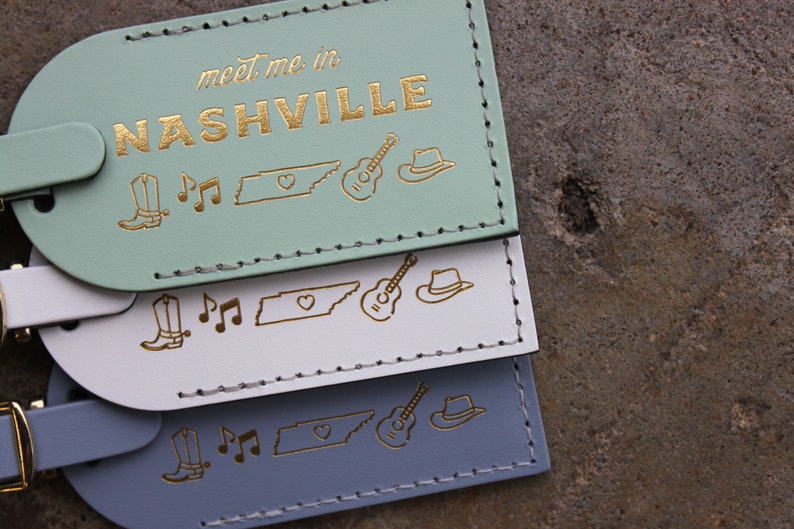May include: Three luggage tags with the text "meet me in NASHVILLE" in gold lettering. The tags are in mint green, white, and light blue. Each tag features a gold outline of a cowboy boot, musical notes, the state of Tennessee, a guitar, and a cowboy hat.