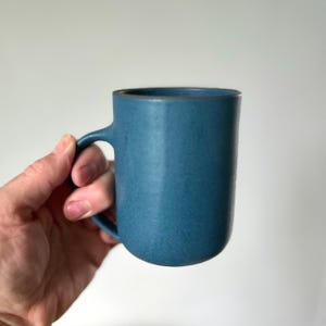 May include: A blue ceramic mug with a rounded handle. The mug has a textured surface and a dark rim. The mug is held by a hand against a white background. The mug is designed for hot beverages.