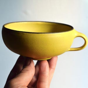 May include: A yellow ceramic teacup with a rounded bowl and a small handle. The rim of the cup has a dark brown trim. The cup is held in a person's hand against a white background. The cup has a textured surface.