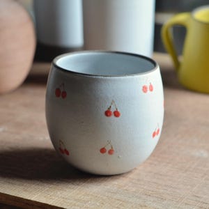 Puede incluir: Una taza de cerámica blanca con forma redondeada, adornada con pequeñas ilustraciones de cerezas rojas. La taza tiene un borde oscuro y está sobre una superficie de madera. Otros artículos de cerámica son visibles en el fondo.