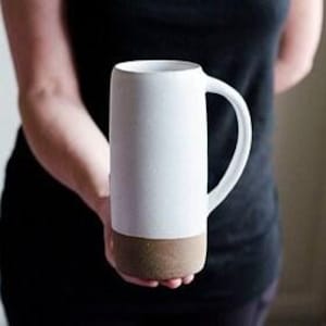 May include: A tall, white ceramic mug with a cylindrical shape and a brown, textured base. The mug has a curved handle and is held by a person wearing a black shirt.