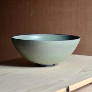 Pode incluir: Uma tigela de cerâmica verde clara com borda e base escuras. A tigela está sobre uma superfície de madeira clara contra um fundo marrom. O design da tigela é simples e elegante, adequado para servir alimentos.