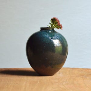 May include: A handcrafted ceramic vase with a rounded body and a narrow neck. The vase features a gradient of colors, transitioning from a deep blue-green at the top to a warm brown at the base. A small sprig of pink flowers adds a touch of color.