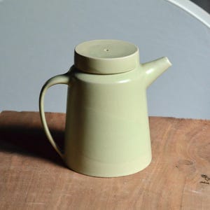 Op de afbeelding: Een lichtgroene keramische theepot met bijpassende deksel. De theepot heeft een gebogen handvat en een tuit. Het deksel heeft een klein gaatje in het midden. De theepot staat op een houten oppervlak.