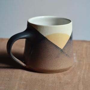May include: A ceramic mug with a black handle and a design featuring a mountain-like pattern in shades of brown, tan, and black. The interior is white, and the mug sits on a wooden surface. This handmade mug is perfect for coffee or tea.
