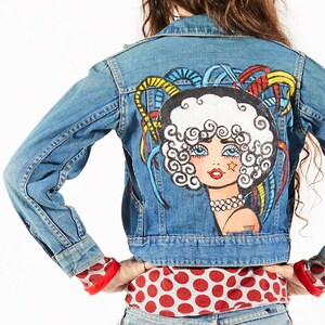 May include: A blue denim jacket with a hand-painted design of a woman with white hair and a colorful background.
