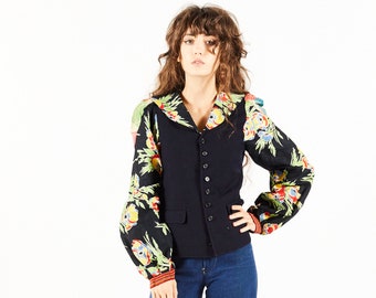 May include: A woman wearing a black vest with a floral print shirt underneath. She is wearing blue jeans and brown platform shoes.