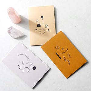 Puede incluir: Tres tarjetas con dibujos abstractos en blanco y negro sobre fondos de colores diferentes. Las tarjetas están sobre una superficie blanca con dos cristales rosas y blancos.