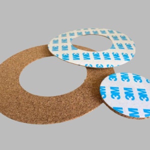 Puede incluir: Tres círculos de corcho con respaldo adhesivo. El círculo más grande es marrón y tiene un agujero en el centro. Los dos círculos más pequeños son blancos con la marca 3M azul.