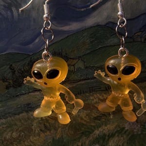 May include: A pair of dangle earrings featuring small, translucent yellow alien figures with black eyes. The alien figures are posed with arms outstretched, and are suspended from silver-colored fish hook ear wires. The earrings are set against a blurred background.