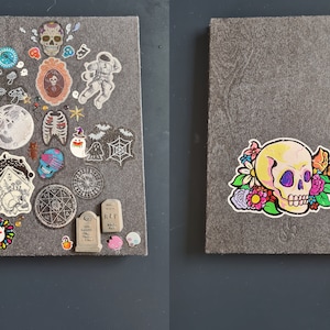 May include: A gray notebook with a red ribbon bookmark. The front cover is decorated with various colorful stickers, including a skull with flowers, a moon, a skeleton, and a ghost. The back cover has a single sticker of a skull with flowers.