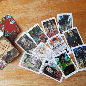 May include: A deck of Villain Clans II: Revenge of the Deck playing cards featuring illustrations of various villains from horror movies. The cards are laid out on a wooden surface. The box for the cards is in the lower left corner of the image. The box is red with black and white illustrations of villains.