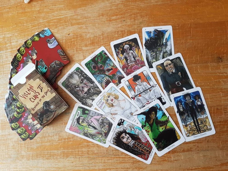 May include: A deck of Villain Clans II: Revenge of the Deck playing cards featuring illustrations of various villains from horror movies. The cards are laid out on a wooden surface. The box for the cards is in the lower left corner of the image. The box is red with black and white illustrations of villains.