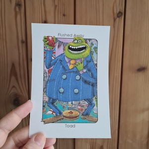 May include: A cartoon illustration of a green toad character from the movie "Flushed Away" wearing a blue suit and holding an apple. The image has the text "Flushed Away" at the top and "Toad" at the bottom.
