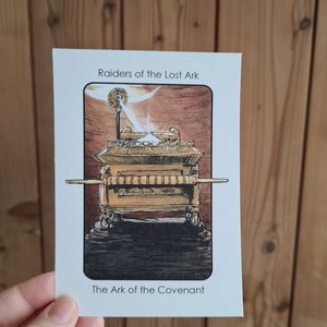 Può includere: Una carta con un'illustrazione dell'Arca dell'Alleanza dal film I predatori dell'arca perduta. L'Arca è un forziere d'oro con un coperchio e due cherubini in cima. La carta ha il testo "I predatori dell'arca perduta" e "L'Arca dell'Alleanza".