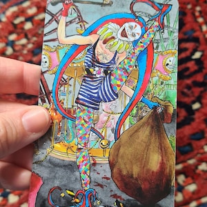 May include: Tarot card illustration of "The Fool" featuring a jester-like figure in colorful attire, including a striped top and patterned leggings. The background depicts a carousel scene with a brown sack. The card's title is at the bottom.