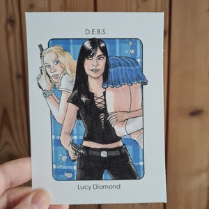 Peut inclure: Une illustration de bande dessinée de deux femmes, l'une avec des cheveux blonds et l'autre avec des cheveux noirs, tenant des armes à feu. La femme aux cheveux noirs porte un haut noir avec un laçage sur le devant et un pantalon noir. La femme aux cheveux blonds porte une chemise blanche et une jupe bleue. L'arrière-plan est bleu avec des diamants blancs. Le texte "D.E.B.S." et "Lucy Diamond" est imprimé sous l'image.