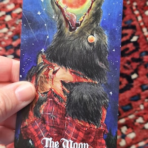 May include: A tarot card illustration depicting a wolf with an open mouth against a night sky with a full moon. The wolf is tearing at a person wearing a red plaid shirt. The card reads "The Moon."