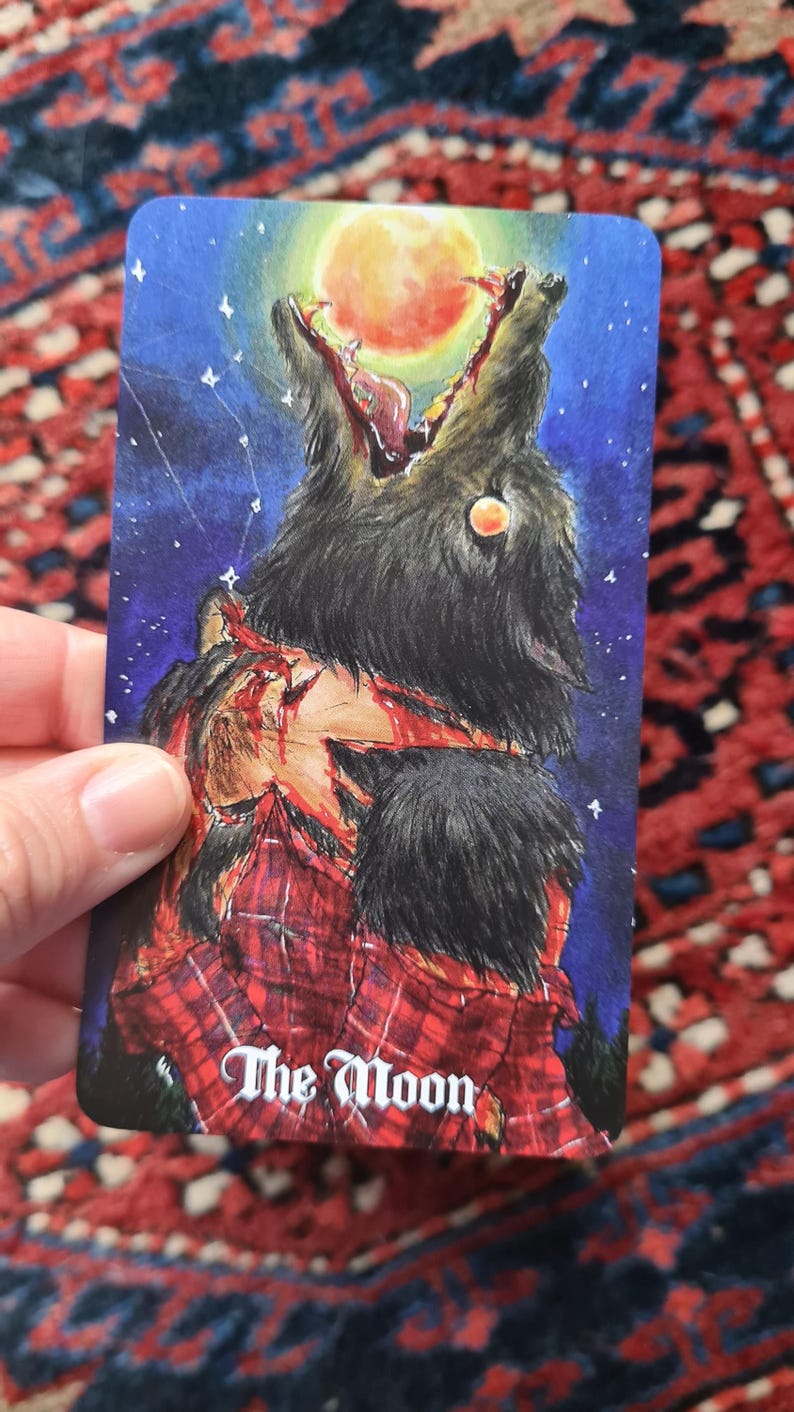 May include: A tarot card illustration depicting a wolf with an open mouth against a night sky with a full moon. The wolf is tearing at a person wearing a red plaid shirt. The card reads "The Moon."