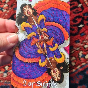 May include: A tarot card featuring a woman in a blue and orange dress holding a sword. The card is labeled "2 of Swords". The woman has dark skin and curly black hair. The background is white.
