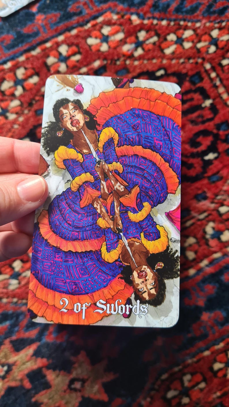 May include: A tarot card featuring a woman in a blue and orange dress holding a sword. The card is labeled "2 of Swords". The woman has dark skin and curly black hair. The background is white.