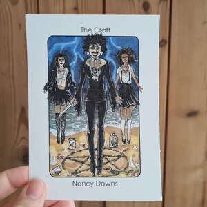 May include: A tarot card with an illustration of three women in black clothing standing in a circle with candles and a pentagram. The card is titled "The Craft" and the artist is credited as "Nancy Downs".