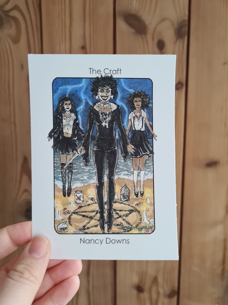 May include: A tarot card with an illustration of three women in black clothing standing in a circle with candles and a pentagram. The card is titled "The Craft" and the artist is credited as "Nancy Downs".