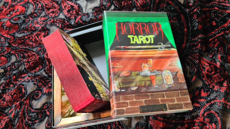 May include: A tarot card deck titled "HORROR TAROT" with a green and red cover. The deck is open, revealing the cards with red edges. The box features a spooky illustration of a room with a brick wall.