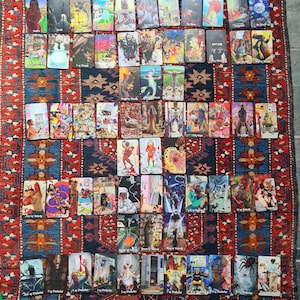May include: A collection of tarot cards, each featuring unique illustrations and titles, arranged on a patterned rug. The cards display a variety of artistic styles and themes, with vibrant colors and detailed imagery. The rug has a red, blue, and cream color scheme.