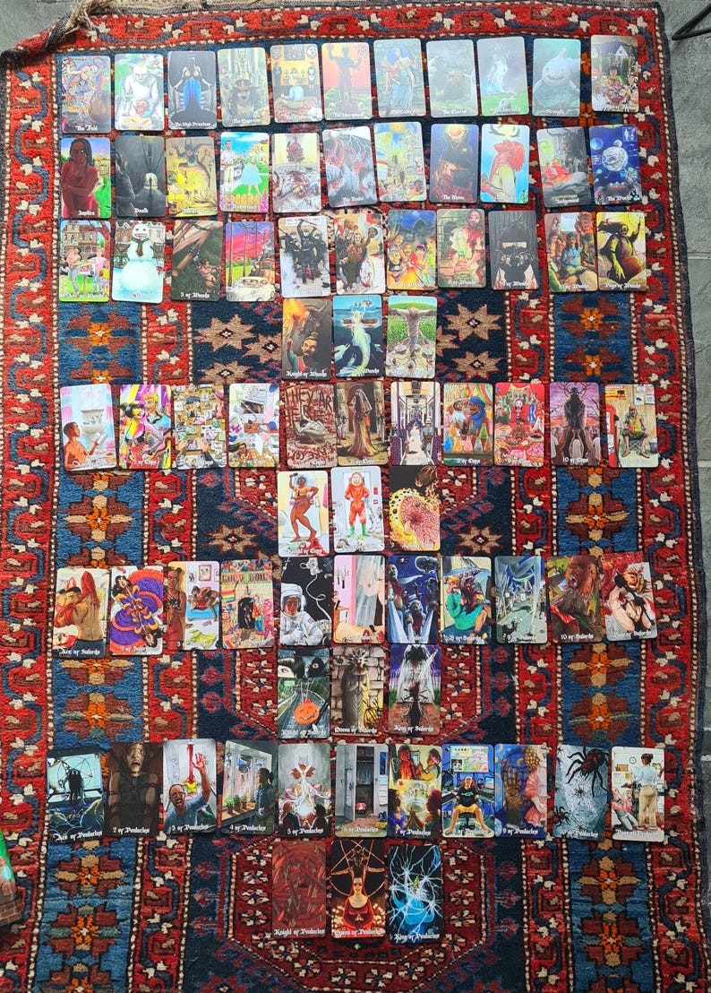 May include: A collection of tarot cards, each featuring unique illustrations and titles, arranged on a patterned rug. The cards display a variety of artistic styles and themes, with vibrant colors and detailed imagery. The rug has a red, blue, and cream color scheme.
