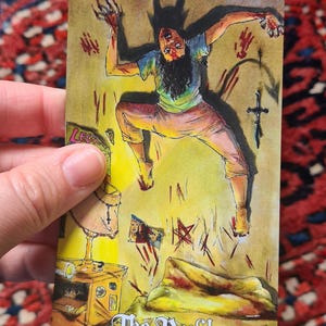 May include: Tarot card illustration featuring a figure with horns and claws, labeled "The Devil." The card depicts a person in a distressed pose, surrounded by symbols and a cross. The background is yellow and the card is held by a hand.