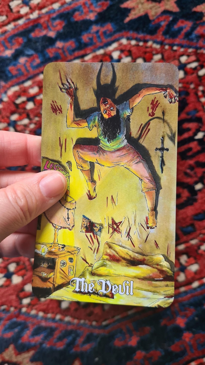 May include: Tarot card illustration featuring a figure with horns and claws, labeled "The Devil." The card depicts a person in a distressed pose, surrounded by symbols and a cross. The background is yellow and the card is held by a hand.