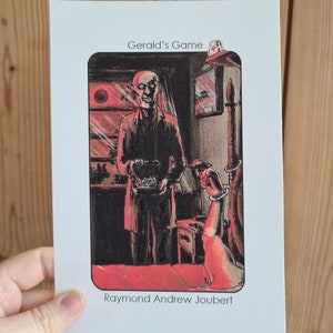 May include: A black and white illustration of a man in a room with a red moon in the window. The man is holding a box and looking at the viewer. The text "Gerald's Game" is at the top of the image and "Raymond Andrew Joubert" is at the bottom.
