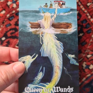 May include: A tarot card illustration featuring a mermaid with a flowing, light-colored tail and hair, emerging from the water. A boat with people is in the background. The card is labeled "Queen of Wands".