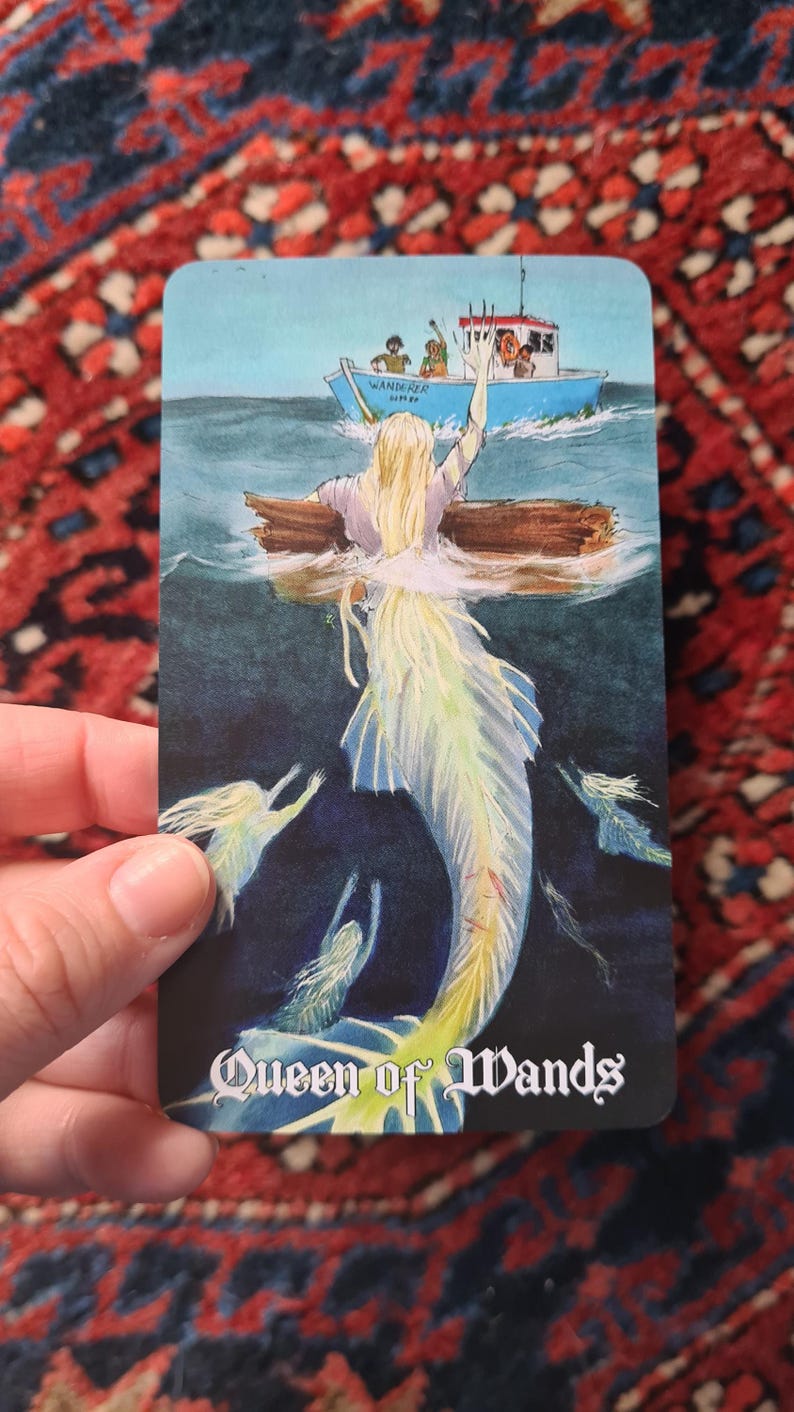 May include: A tarot card illustration featuring a mermaid with a flowing, light-colored tail and hair, emerging from the water. A boat with people is in the background. The card is labeled "Queen of Wands".