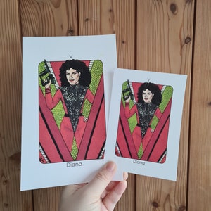 Puede incluir: Dos impresiones art&iacute;sticas blancas que representan a una mujer con un atuendo rojo y negro sosteniendo un arma. Las impresiones tienen un fondo rojo y verde con el nombre "Diana" en la parte inferior. Las impresiones son de diferentes tama&ntilde;os.