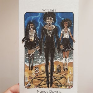 May include: A tarot card illustration depicting three women dressed in black, standing in a circle with candles and a pentagram. The card is titled "Witches" and signed "Nancy Downs".